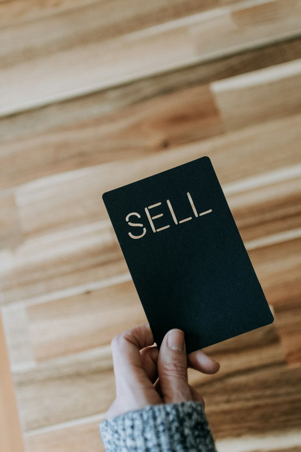 An individual holds a black card with the word 'SELL' cut out in bold letters. The background is a wooden surface. The image evokes themes of business and trading. — Credit: Photo by Kelly Sikkema on Unsplash