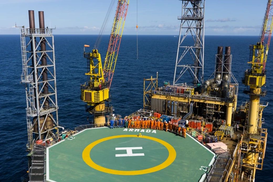 An aerial view of an offshore oil drilling platform, showcasing workers in orange safety suits gathered near a helipad. The surrounding ocean reflects clear skies and horizons, highlighting the industrial scale of the operation.