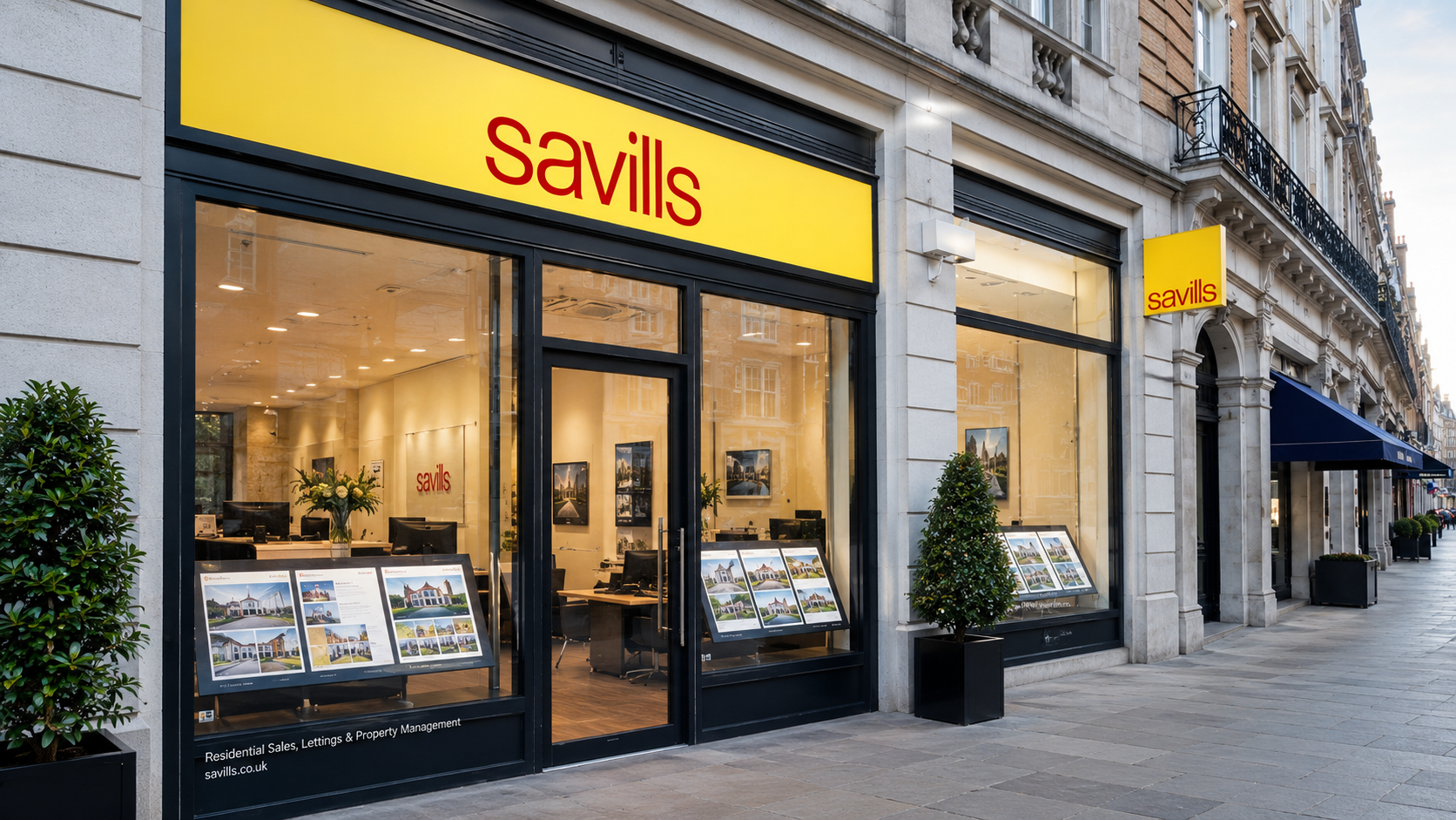 The image shows the exterior of a Savills real estate office located on a city street. The storefront features large windows displaying framed property photographs and a bright yellow sign with the company name.