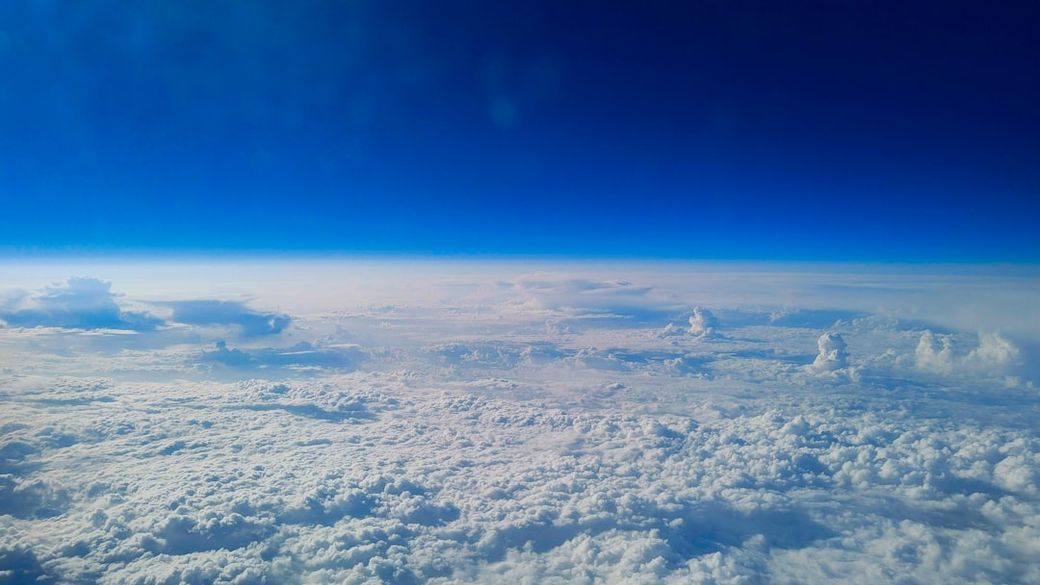 An aerial view of expansive fluffy clouds under a clear blue sky, capturing the vastness of the atmosphere. The scene portrays a serene and calm environment above the cloud layer. — Credit: Photo by Alexander Gluschenko on Unsplash