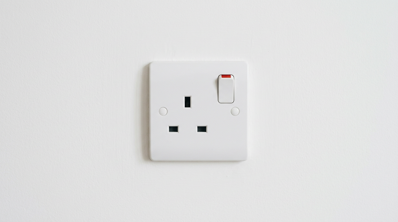 The image shows a close-up view of a standard UK three-pin electrical socket mounted on a clean, white wall. The socket is switched on, indicated by a visible red rocker switch, while the three rectangular slots remain empty, suggesting availability and potential energy awaits demand.