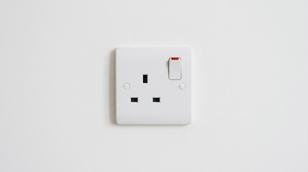 The image shows a close-up view of a standard UK three-pin electrical socket mounted on a clean, white wall. The socket is switched on, indicated by a visible red rocker switch, while the three rectangular slots remain empty, suggesting availability and potential energy awaits demand.