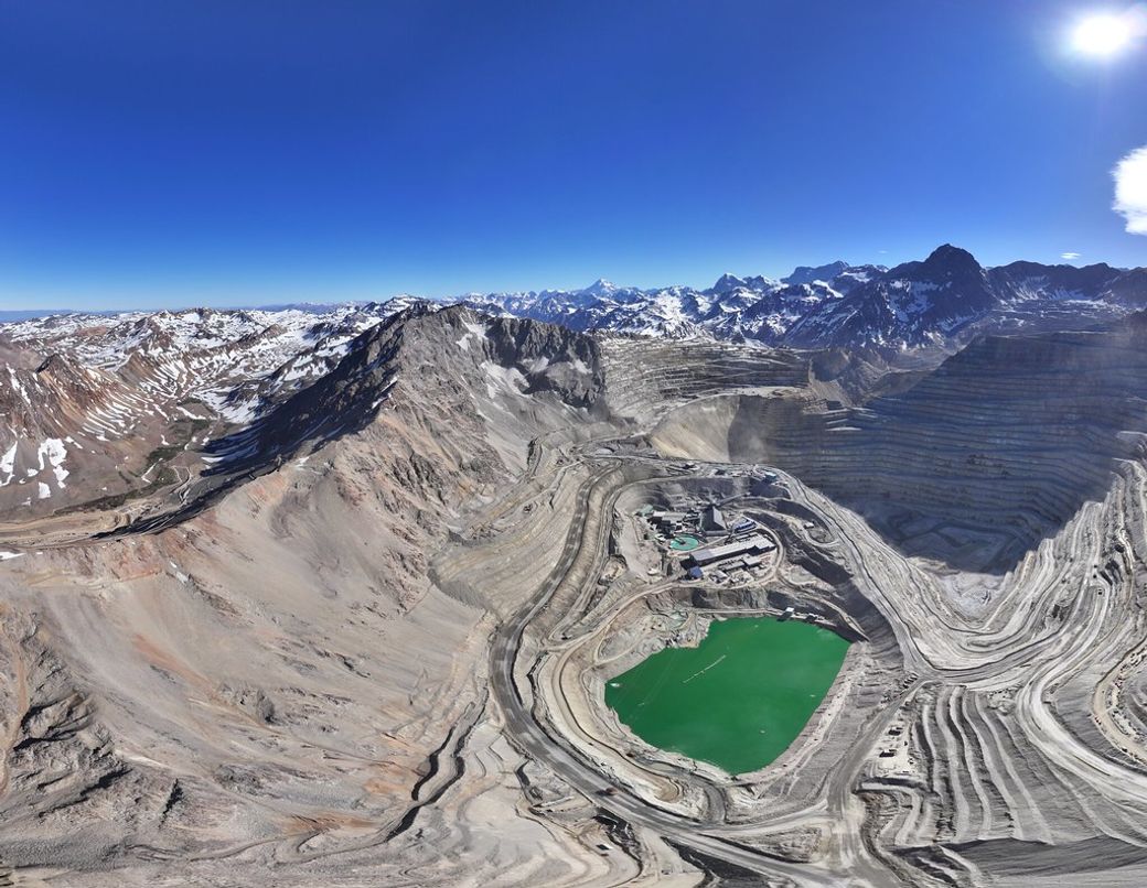 A panoramic view of a large open-pit mining operation surrounded by rugged mountains. A vibrant green body of water reflects the clear blue sky, highlighting the environmental impact of the mining activity.