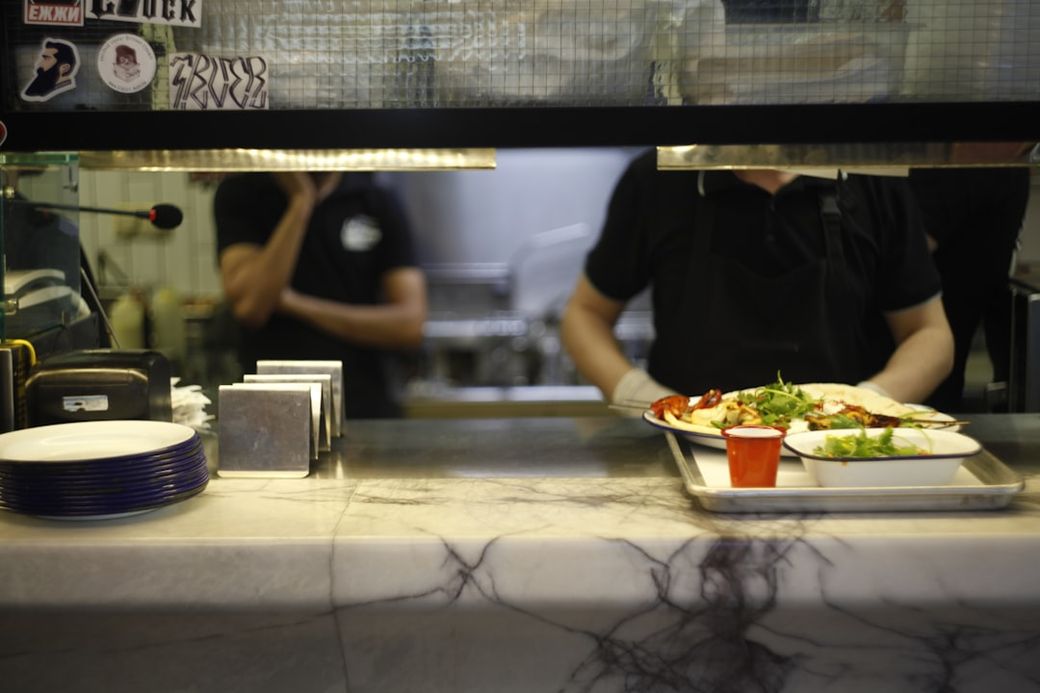 A bustling kitchen scene where a chef is presenting a plate of food, indicating a busy restaurant environment. The countertop is adorned with various kitchen tools and plates, emphasizing the culinary aspect. — Credit: Photo by Anton Murygin on Unsplash