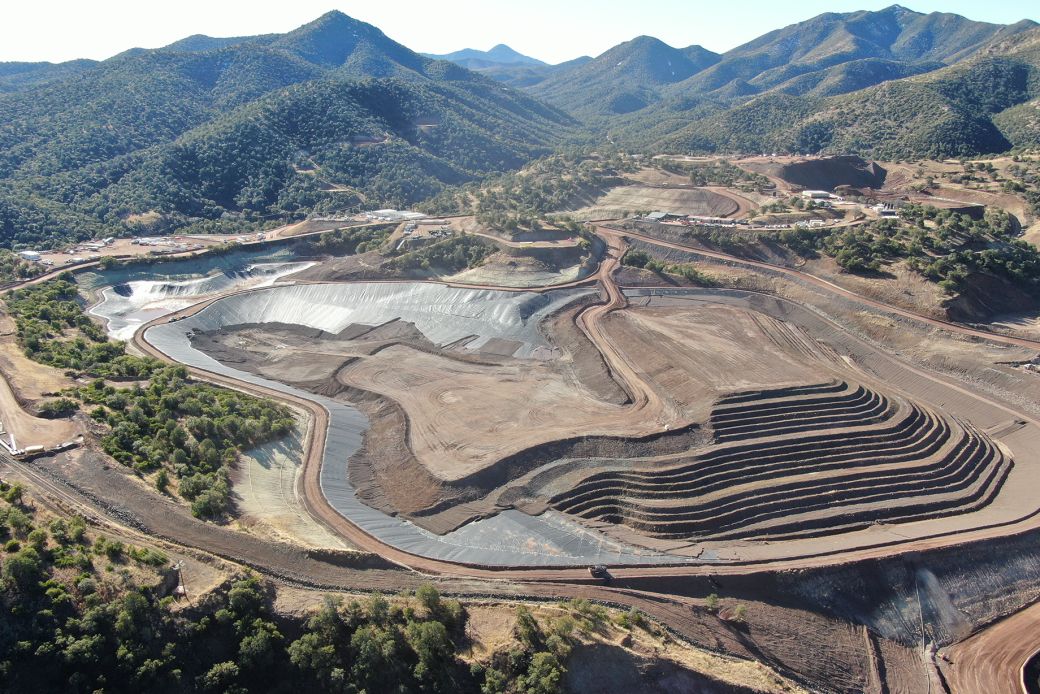 The image shows an aerial view of a mining operation situated in a mountainous area. The landscape features terraced earthworks and various stages of excavation, highlighting the extensive alteration of the terrain for mineral extraction.