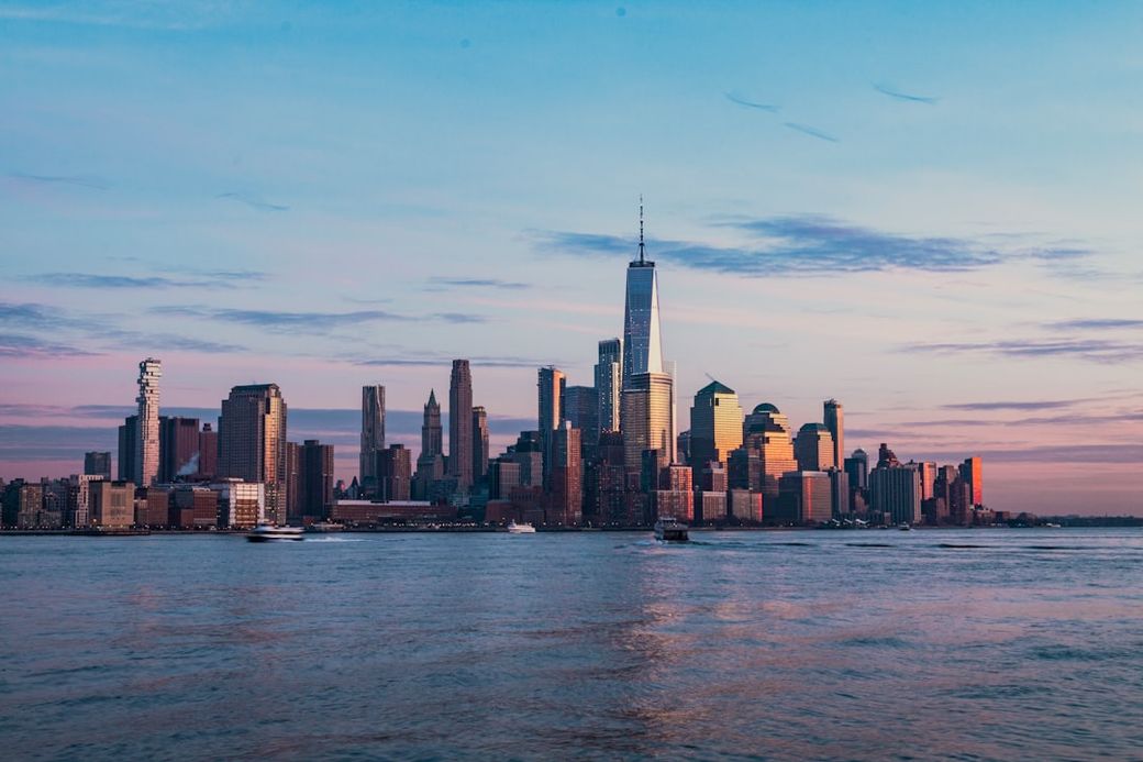 A panoramic view of the New York City skyline during sunset, showcasing the One World Trade Center prominently among other skyscrapers. The scene captures the tranquil waters reflecting the warm hues of the sky. — Credit: Photo by Mike Chavarri on Unsplash