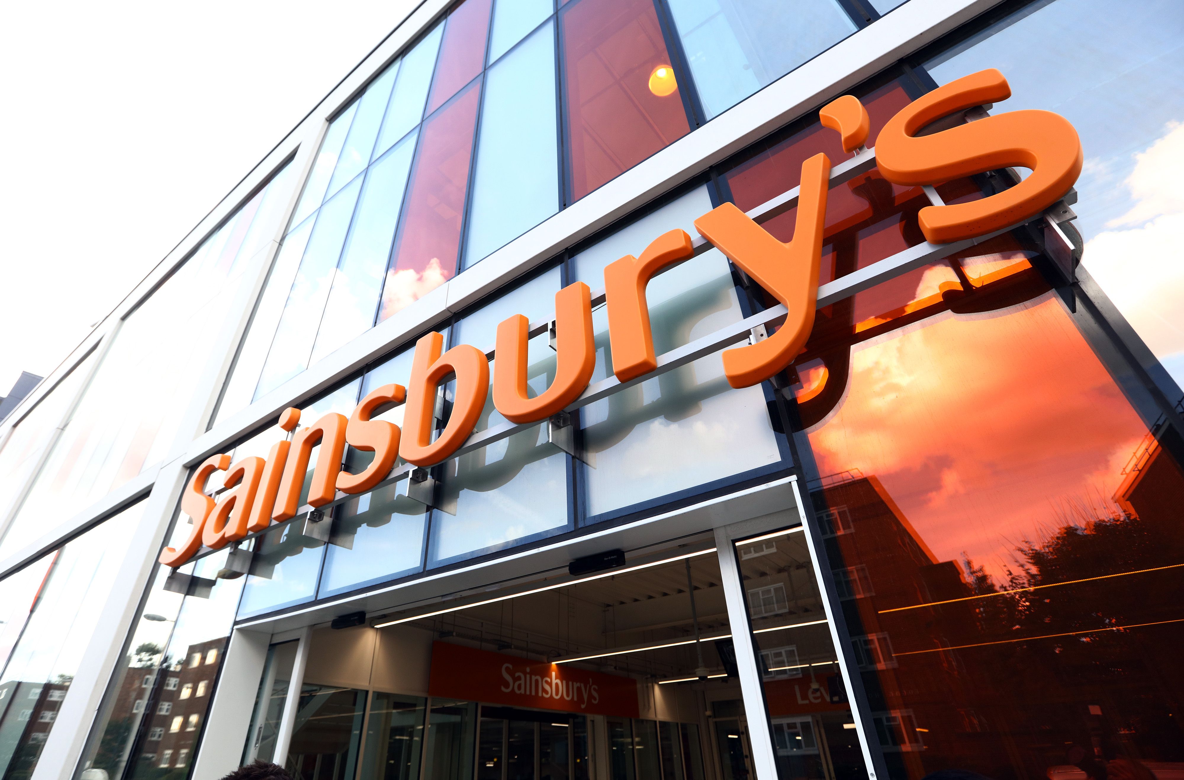 The image showcases the exterior of a Sainsbury's store, featuring large glass windows reflecting the sky. The prominent orange signage highlights the brand name, making it clear that this is a retail grocery location.