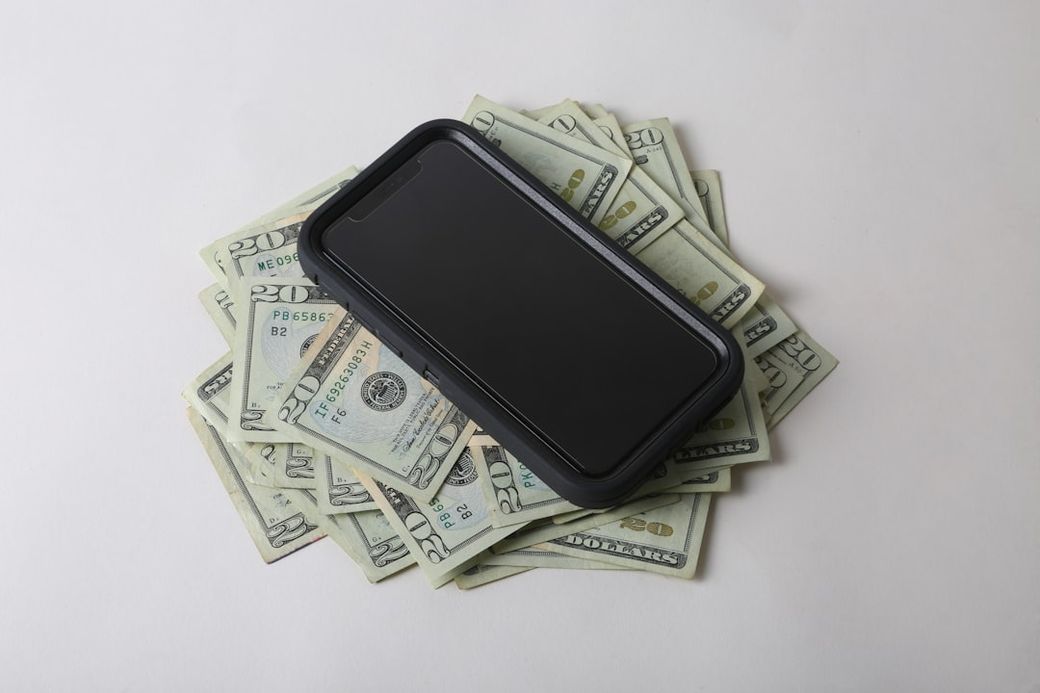The image features a smartphone resting on a pile of cash, specifically U.S. dollar bills. The contrasting elements of technology and currency suggest themes of finance and modern commerce. — Credit: Photo by Giorgio Trovato on Unsplash