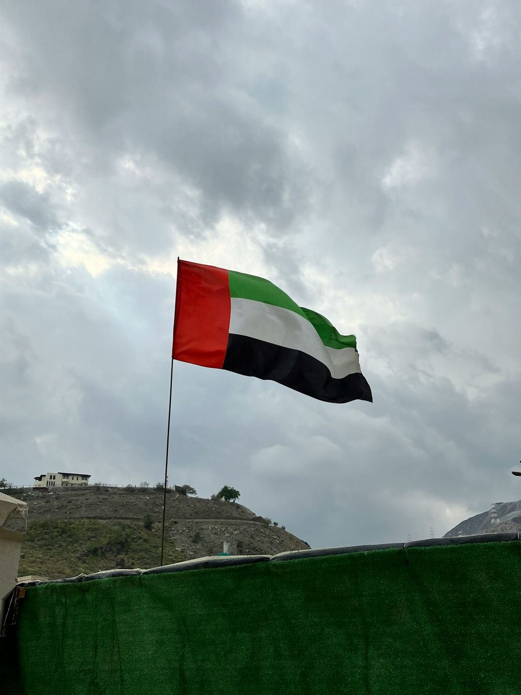 The image features the flag of the United Arab Emirates prominently displayed against a cloudy sky. The background shows a hillside with distinguishable structures. — Credit: Photo by Yegor Gulido on Unsplash