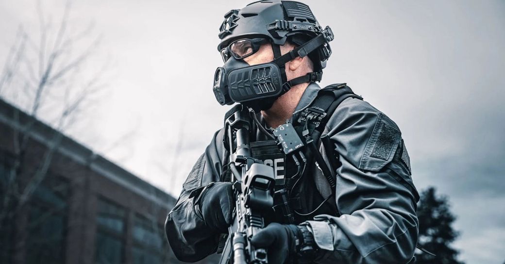 The image depicts an armed tactical officer in full gear, standing in a ready position. The officer wears a helmet with a face mask and is holding a firearm, suggesting a law enforcement or military context.
