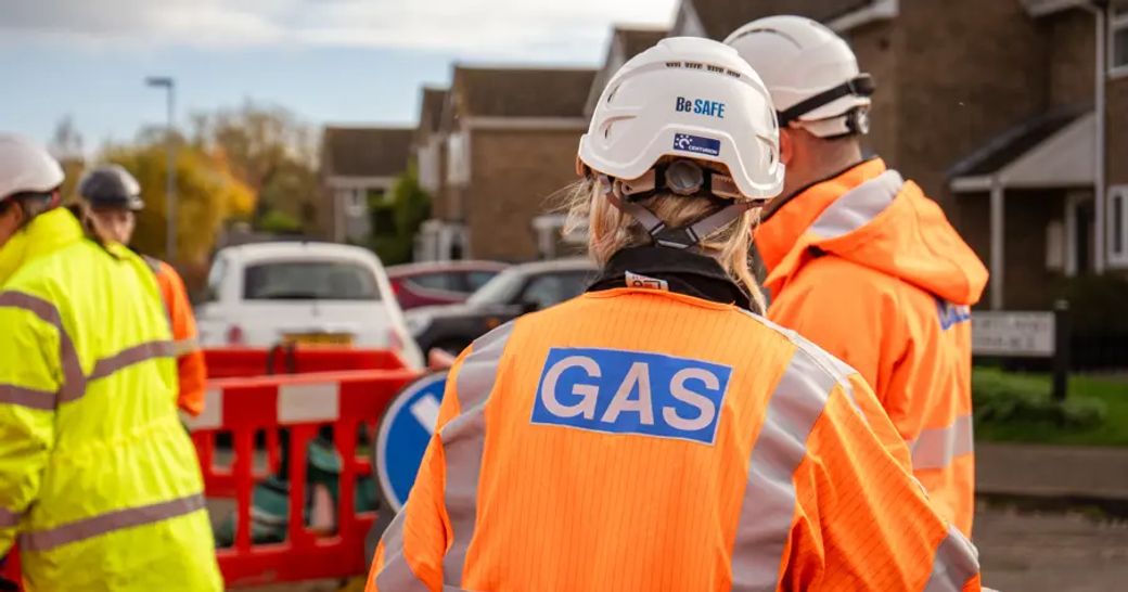 Workers in high-visibility gear and helmets are conducting operations in a residential area. One worker has a visible 'GAS' label on their jacket, indicating a focus on gas safety and services.