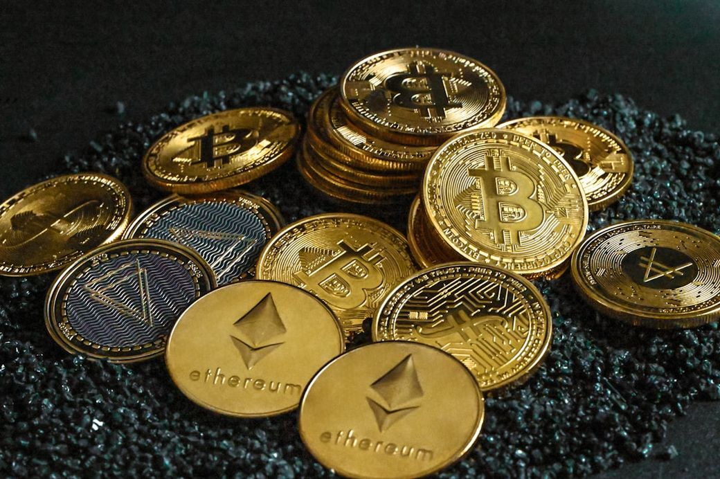 The image features a collection of various cryptocurrency coins, including Bitcoin and Ethereum, laid out on a dark surface. The coins are shiny and reflect light, enhancing their visual appeal. — Credit: Photo by Traxer on Unsplash