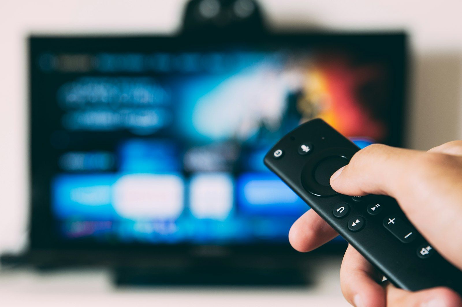 An individual is holding a remote control in front of a television displaying a colorful interface. The screen shows various icons and options commonly associated with streaming platforms.