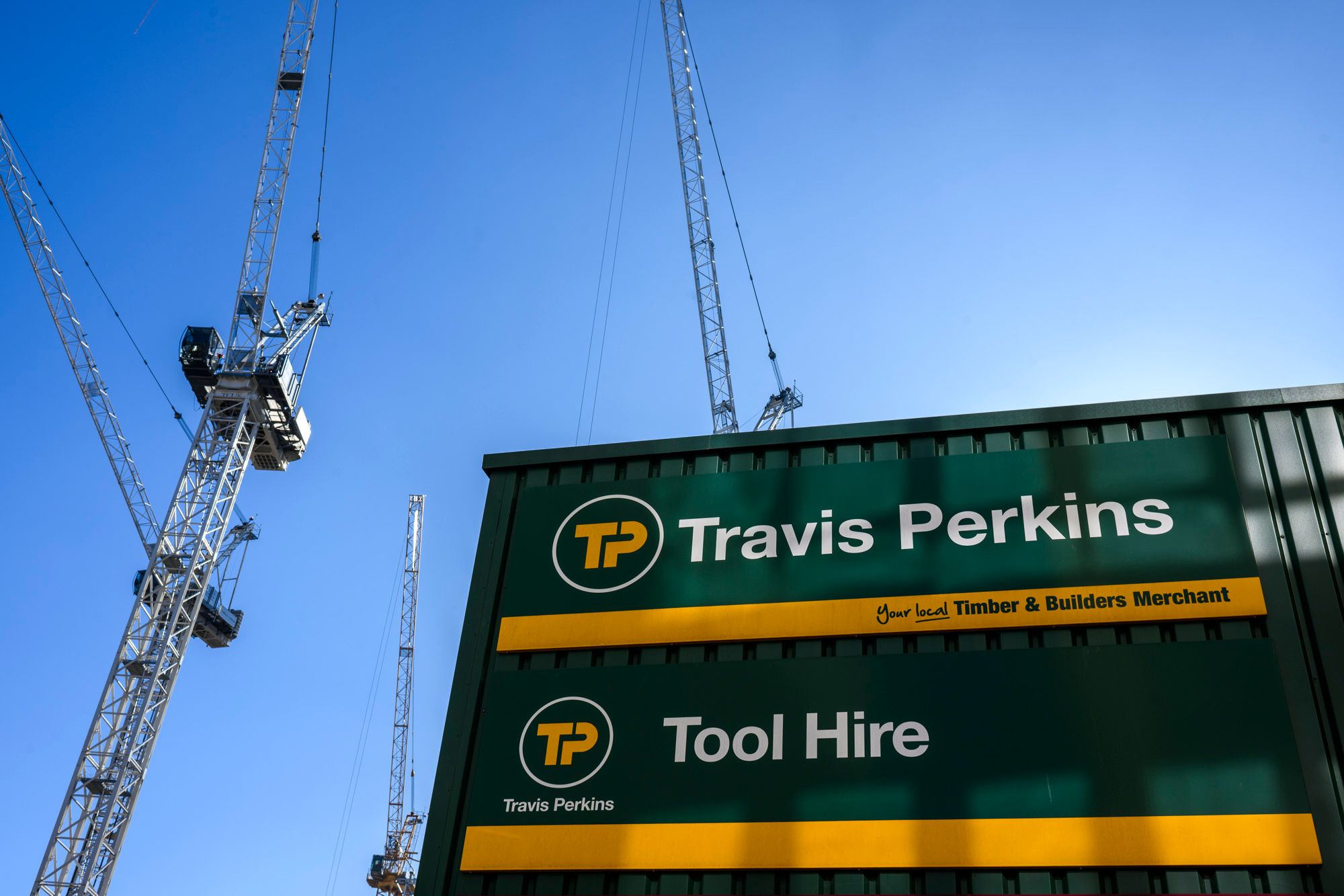 The image showcases a construction site featuring multiple cranes against a clear blue sky. Prominently displayed is a green container with signage for 'Travis Perkins', a well-known builders' merchant, and 'Tool Hire'.