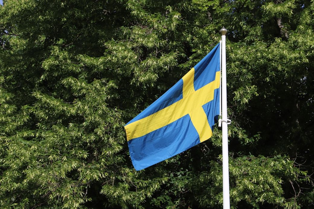 The image shows the national flag of Sweden waving in the wind against a backdrop of lush green trees. The flag features a blue field with a yellow Scandinavian cross. — Credit: Photo by Christer Lässman on Unsplash