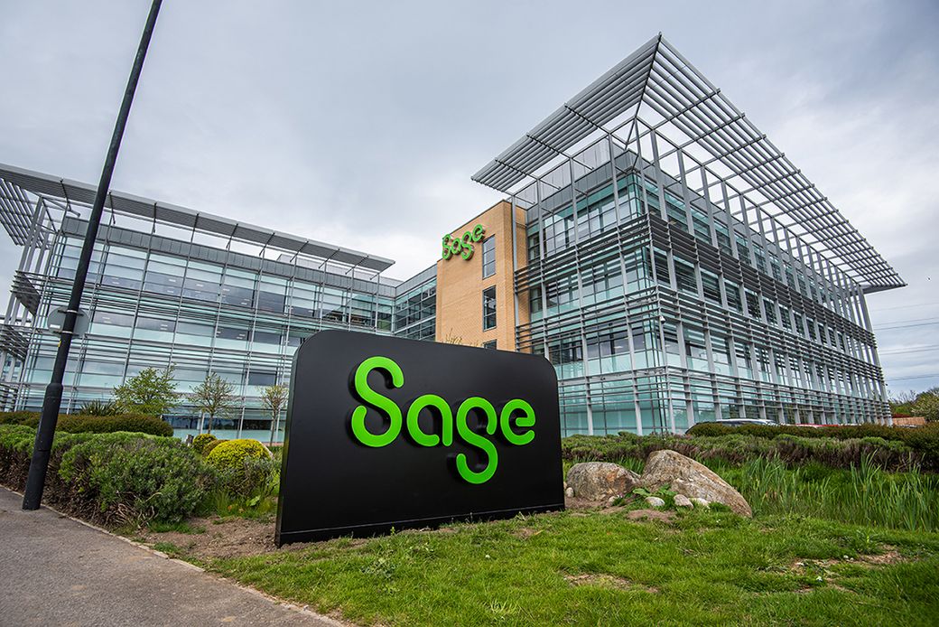 The image shows the modern exterior of a corporate office building belonging to Sage, a company known for software solutions. The building features large glass windows and a prominent sign displaying the Sage logo.