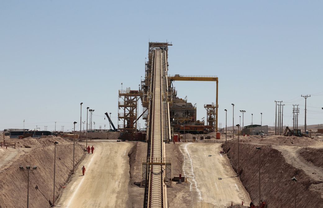 The image depicts an industrial mining site featuring a long conveyor belt system that transports materials. The scene is set in a dry, mountainous area with several workers visible in orange uniforms.