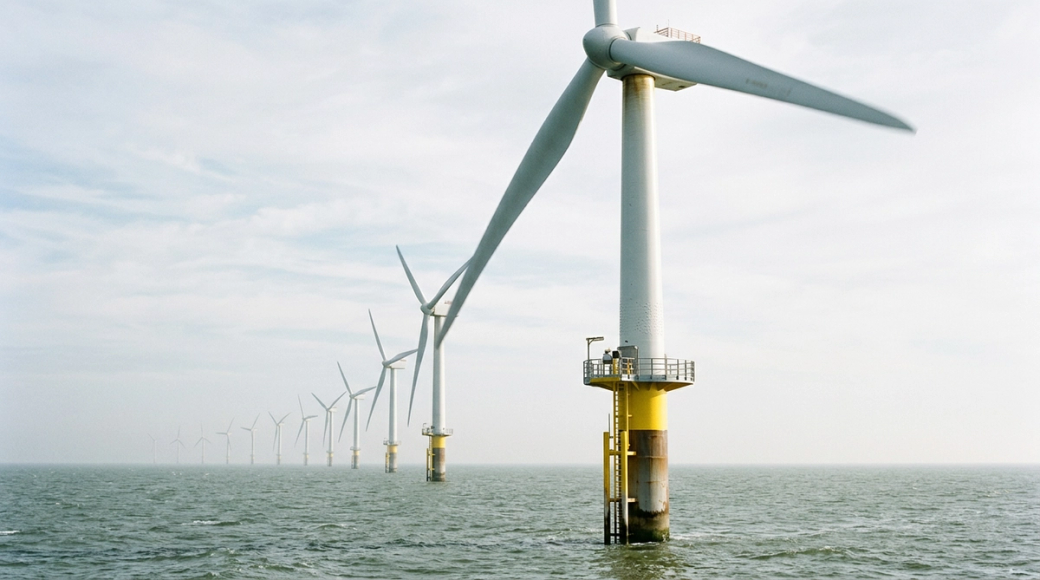 The image depicts a large-scale offshore wind farm with a prominent turbine in the foreground, featuring a monopile foundation rising above the grey-green sea. A series of turbines extends into the distance, creating a sense of depth as they fade into a soft atmospheric haze, all captured under cool marine daylight.