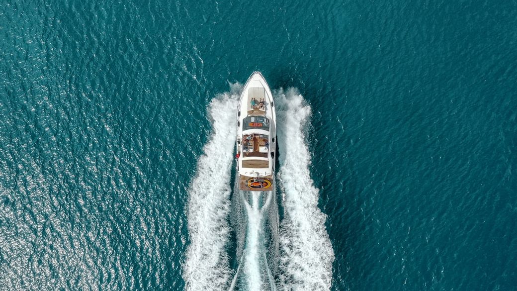 An aerial view of a speedboat moving through turquoise waters, leaving white wake trails behind. The boat is equipped with various recreational items and passengers enjoying the ride. — Credit: Photo by Carles Rabada on Unsplash