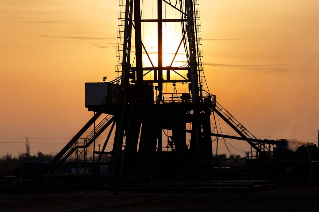 The image depicts a silhouetted oil drilling rig against a sunset, highlighting the structure's intricate details and shadows. The warm orange and yellow hues of the sky create a dramatic backdrop, emphasizing the energy sector's operations. — Credit: Photo by WORKSITE Ltd. on Unsplash