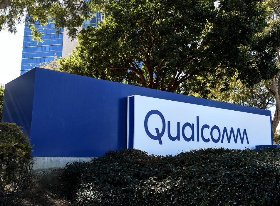 The image features the sign of Qualcomm, prominently displayed in front of a modern building surrounded by trees and shrubs. The sign, which is blue and white, indicates the presence of the technology company in a corporate setting.