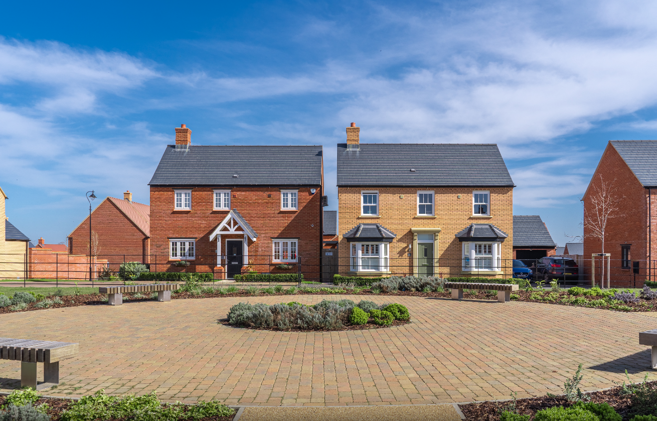 The image depicts a residential area featuring two houses on either side of a circular paved space. The sunny day provides clear visibility of the modern architecture and landscaping.