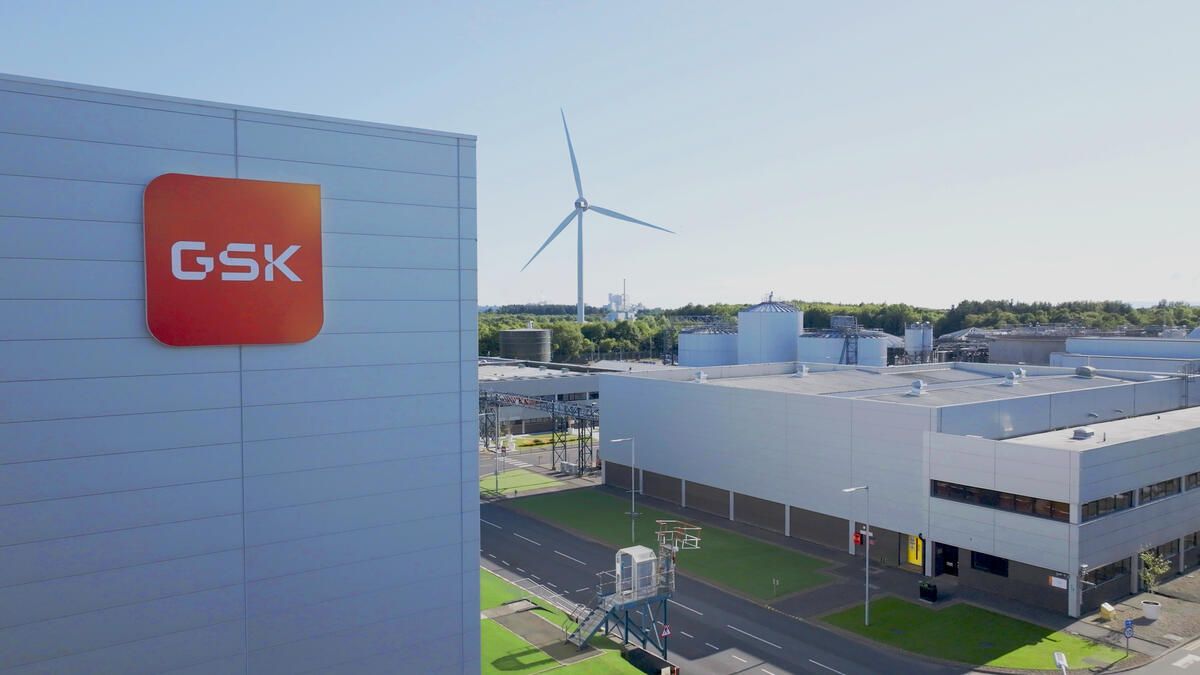 The image shows a modern pharmaceutical manufacturing facility with a prominent GSK logo on the building. A wind turbine is visible in the background, emphasizing the site's commitment to sustainability.