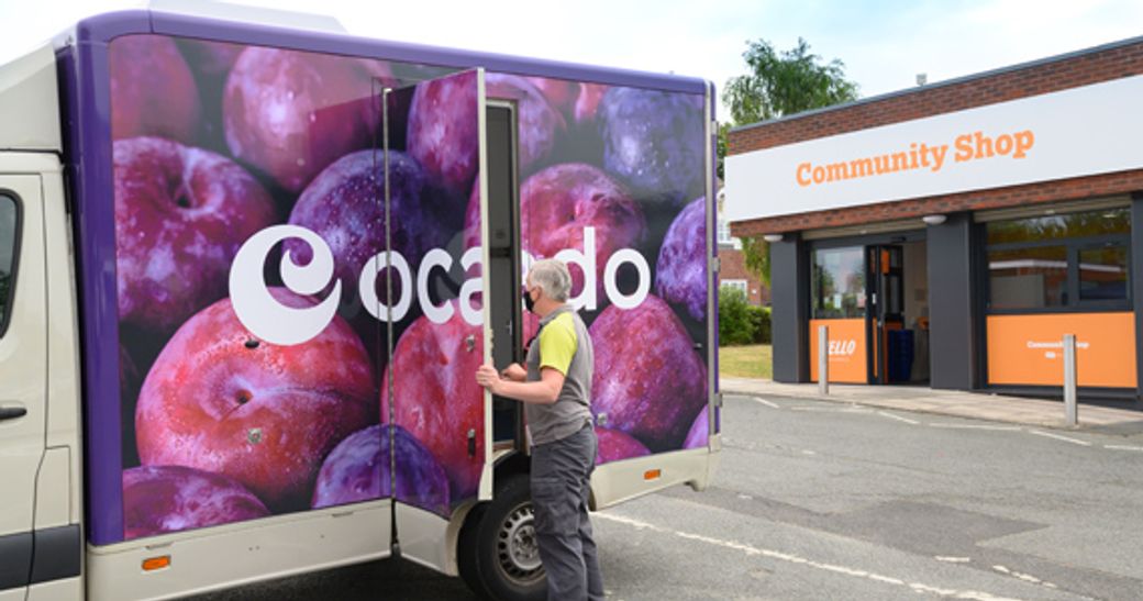 A delivery truck with a large advertisement featuring colorful plums is parked outside a community shop. A person is seen opening the truck's rear door, indicating a delivery or restocking activity.