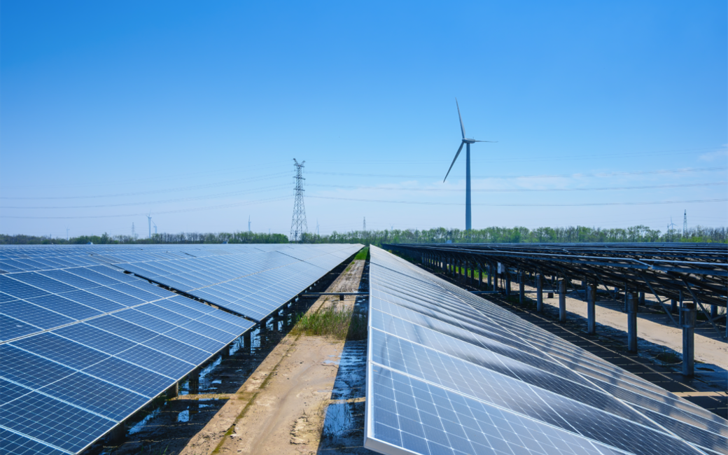 The image showcases a renewable energy site featuring rows of solar panels with a wind turbine in the background under a clear blue sky. The setting emphasizes the integration of solar and wind energy technologies.