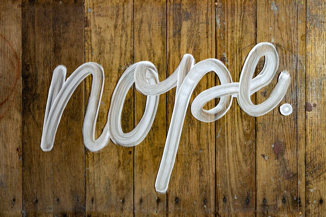 The image features the word 'nope' written in a fluid, white brushstroke style on a textured wooden surface. The composition highlights the contrast between the bright lettering and the warm tones of the wood beneath.
