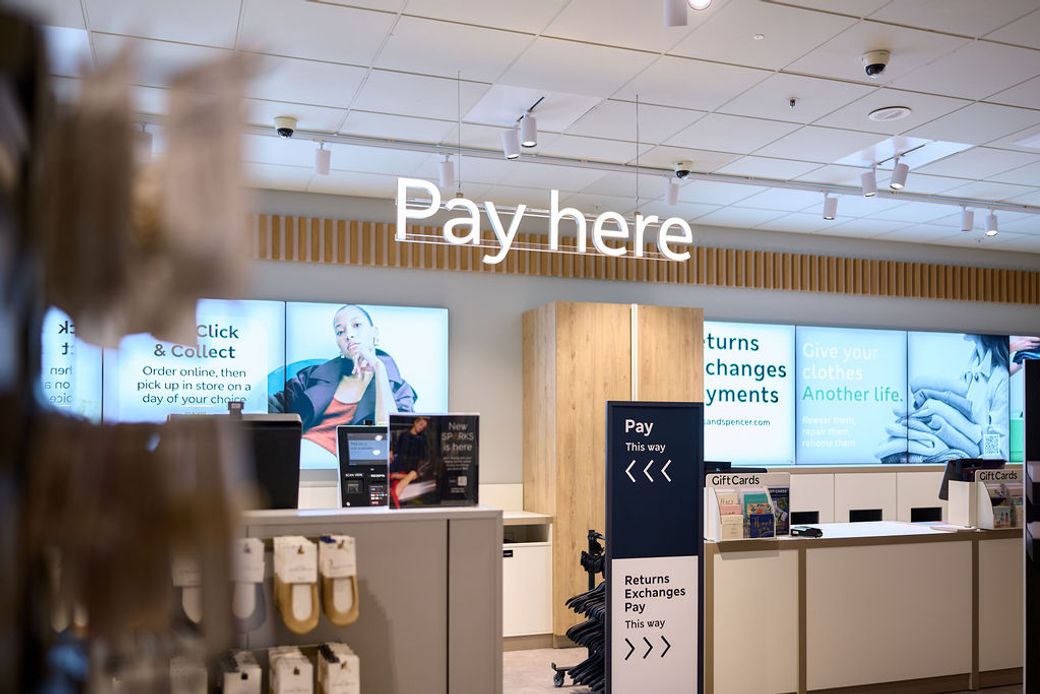 The image shows a retail store checkout area with clear signage indicating payment and returns. A customer is visible in the background, interacting with the store's offerings.