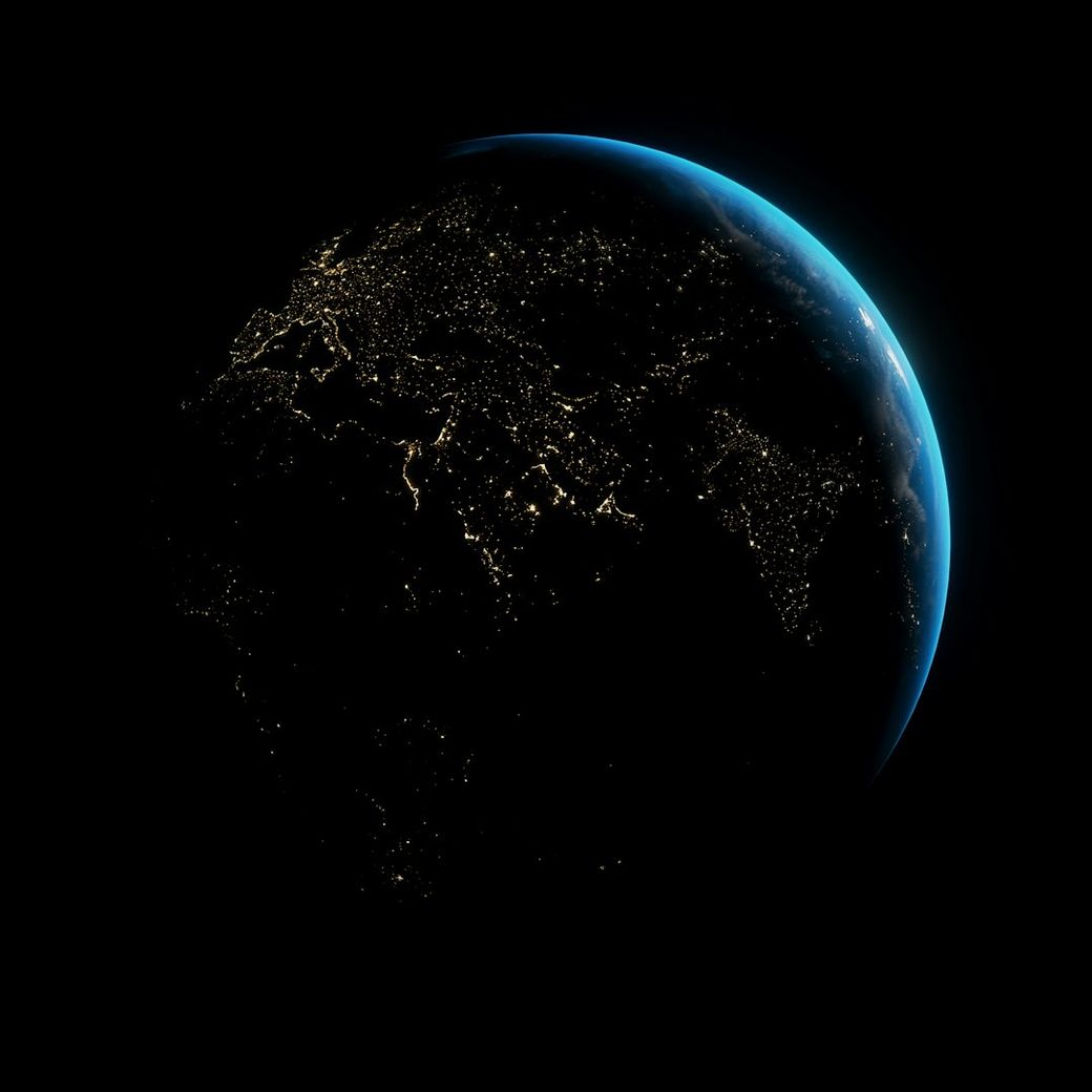 The image shows a nighttime view of Earth, highlighting the illuminated areas indicative of human population and urban development. The planet is depicted in a dark space environment, with visible outlines of continents and coastlines. — Credit: Photo by Anirudh on Unsplash