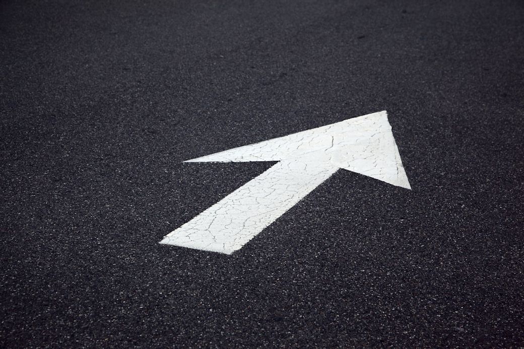 The image shows a cracked white arrow painted on a dark asphalt surface, indicating direction. The arrow points straight ahead, emphasizing guidance on the pavement. — Credit: Photo by Shane on Unsplash