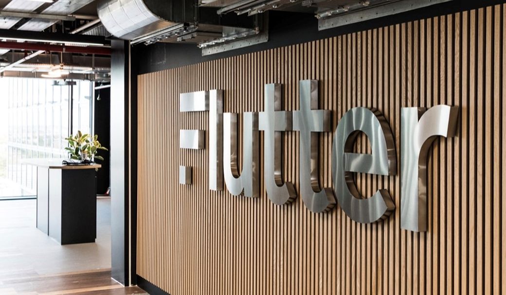 The image shows the interior of an office space featuring a prominent wall with the brand name 'Flutter' in large metallic letters. The setting is modern and minimalist, with wooden paneling and a glimpse of greenery in the background.