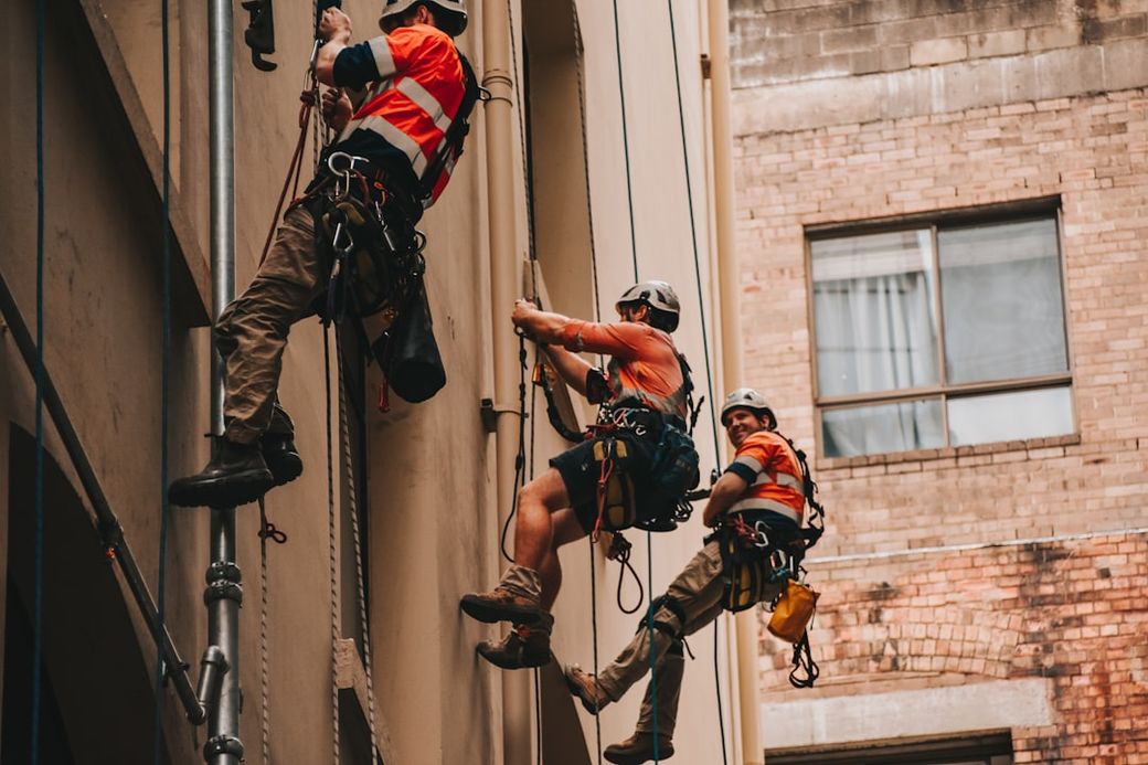 The image depicts three workers in safety gear rappelling down the side of a building. They are secured with harnesses and ropes, performing maintenance or inspection work on the exterior. — Credit: Photo by Dean Bennett on Unsplash
