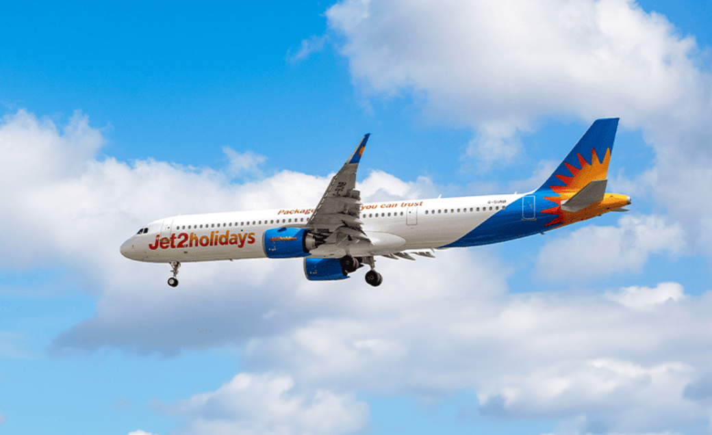 A Jet2holidays airplane is flying against a backdrop of blue skies and fluffy clouds. The aircraft is captured in mid-flight, showcasing its vibrant design and colors.