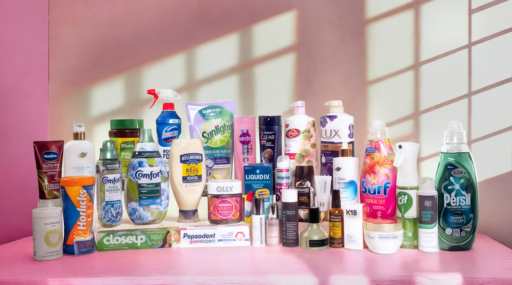 The image showcases a diverse range of personal care and cleaning products arranged against a pink backdrop, highlighting various brands and packaging designs. Sunlight and shadow patterns create a visually engaging atmosphere, enhancing the product display.