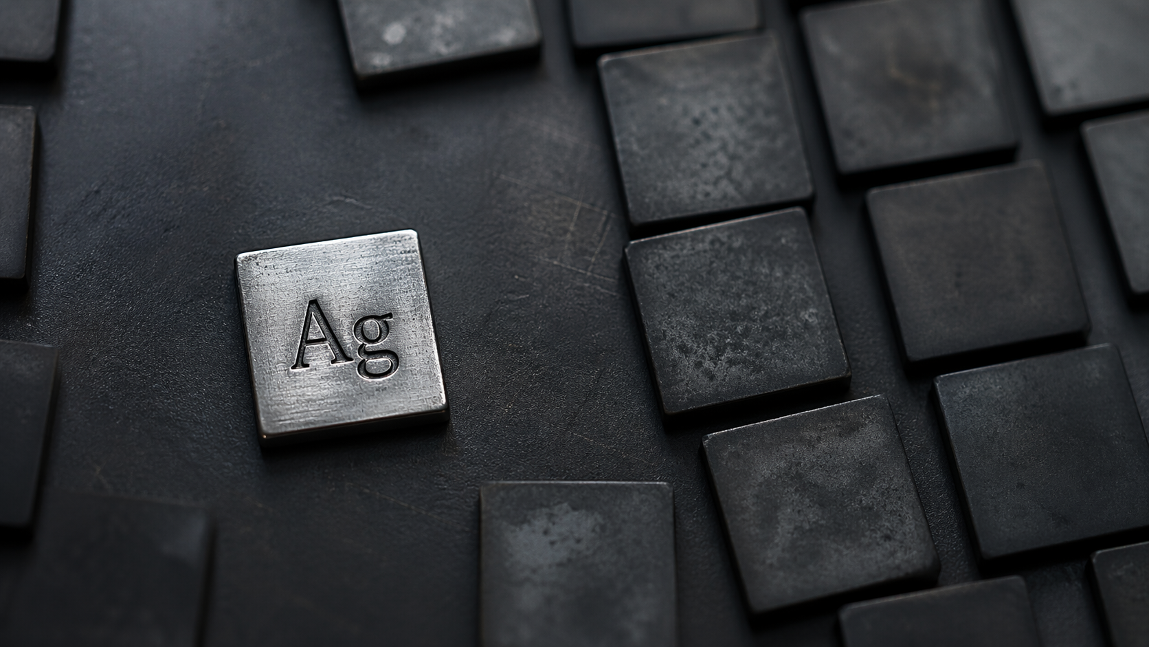 The image features a metal square engraved with the symbol 'Ag', representing silver, placed among other similarly shaped dark metal squares. The focus is on the contrast between the shiny silver square and the matte black surrounding squares.