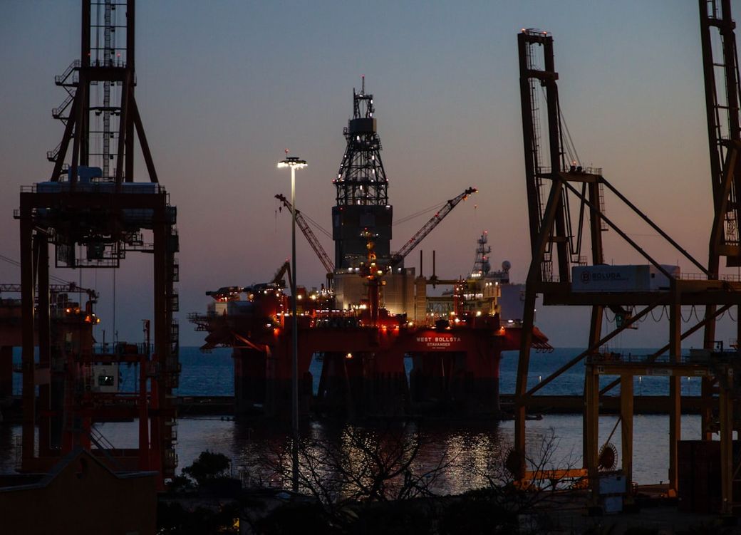 An oil rig is illuminated at dusk, positioned in the sea with cranes in the foreground. The setting sun creates a gradient of colors in the sky, highlighting the industrial activity. — Credit: Photo by Maria Lupan on Unsplash