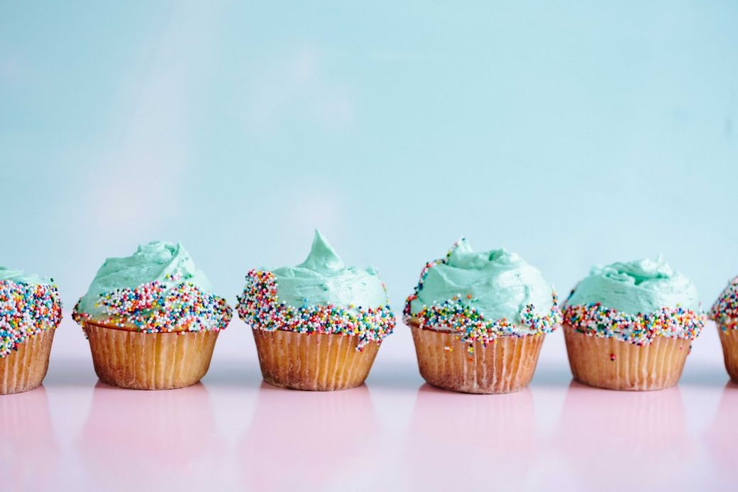A row of decorated cupcakes with light blue frosting and colorful sprinkles are displayed on a pink surface. The backdrop features a soft, pastel blue gradient. — Credit: Photo by Brooke Lark on Unsplash