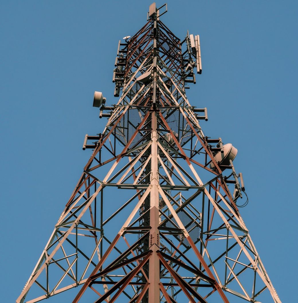 The image features a telecommunications tower viewed from below, showcasing its structure against a clear blue sky. Various antennas and equipment are installed at different heights, indicating its purpose in mobile communication. — Credit: Photo by Ahnaf Piash on Unsplash