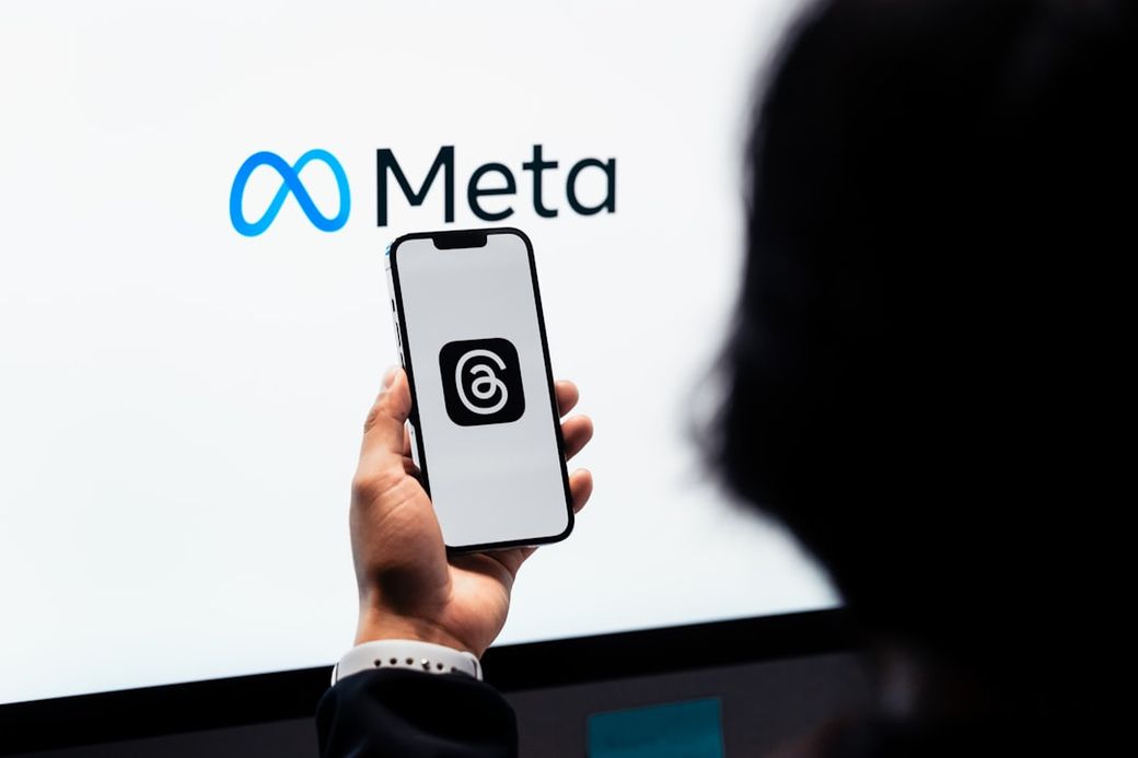 A person holding a smartphone displaying the logo of the Threads app, with the Meta logo visible in the background. The scene suggests a technology-focused environment, possibly a demonstration or event. — Credit: Photo by Julio Lopez on Unsplash