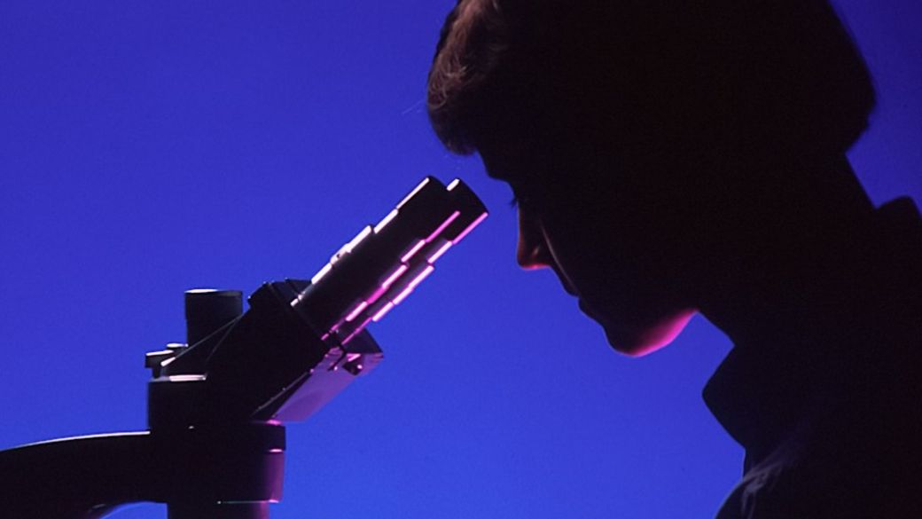 A profile of a female pathologist viewing a histological slide while sitting at a microscope. This dramatic shot has a blue background, with the microscope and the pathologist's features highlighted with magenta. Light is seen passing through the microscope's condensor to the objectives. — Credit: Photo by National Cancer Institute on Unsplash