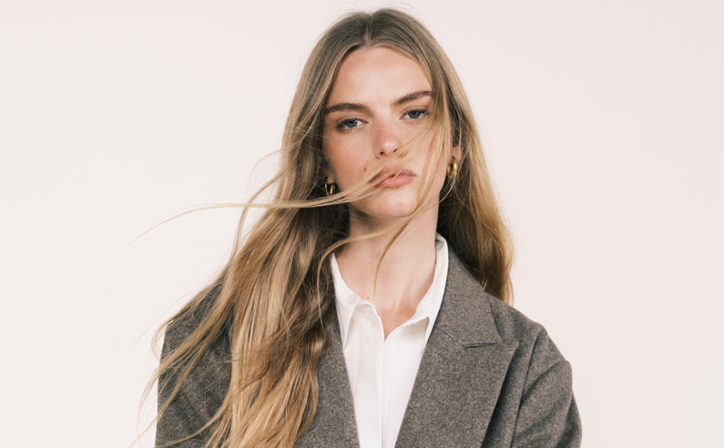 A woman with long hair poses against a neutral background, wearing a stylish blazer over a white shirt. The image captures her serious expression and fashion-forward look.