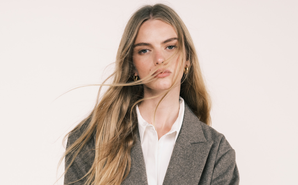 A woman with long hair poses against a neutral background, wearing a stylish blazer over a white shirt. The image captures her serious expression and fashion-forward look.