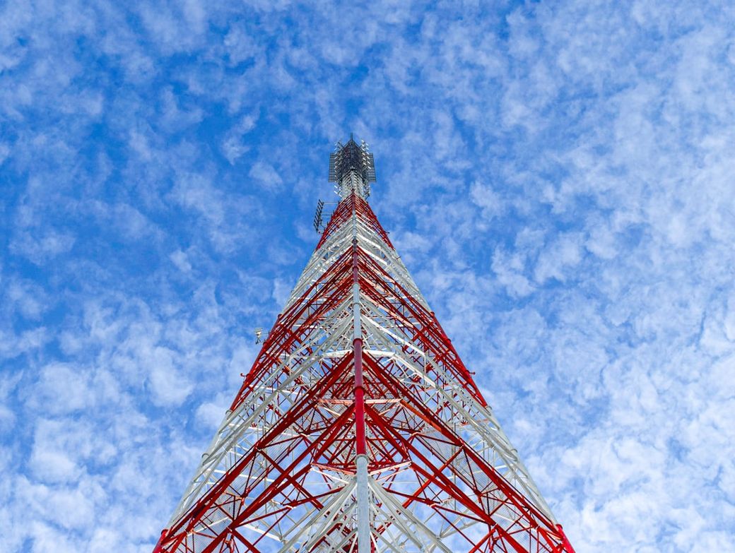 The image shows a tall red and white telecommunications tower reaching towards a blue sky adorned with scattered clouds. The perspective is from the ground looking up, emphasizing the height and structure of the tower. — Credit: Photo by Nopparuj Lamaikul on Unsplash