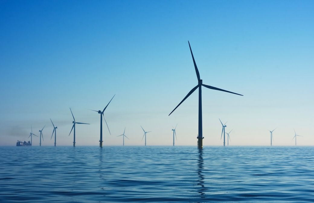 The image depicts a serene view of multiple wind turbines situated offshore in the ocean, surrounded by calm waters under a clear blue sky. The scene showcases renewable energy technology and its integration into the marine environment. — Credit: Photo by Nicholas Doherty on Unsplash