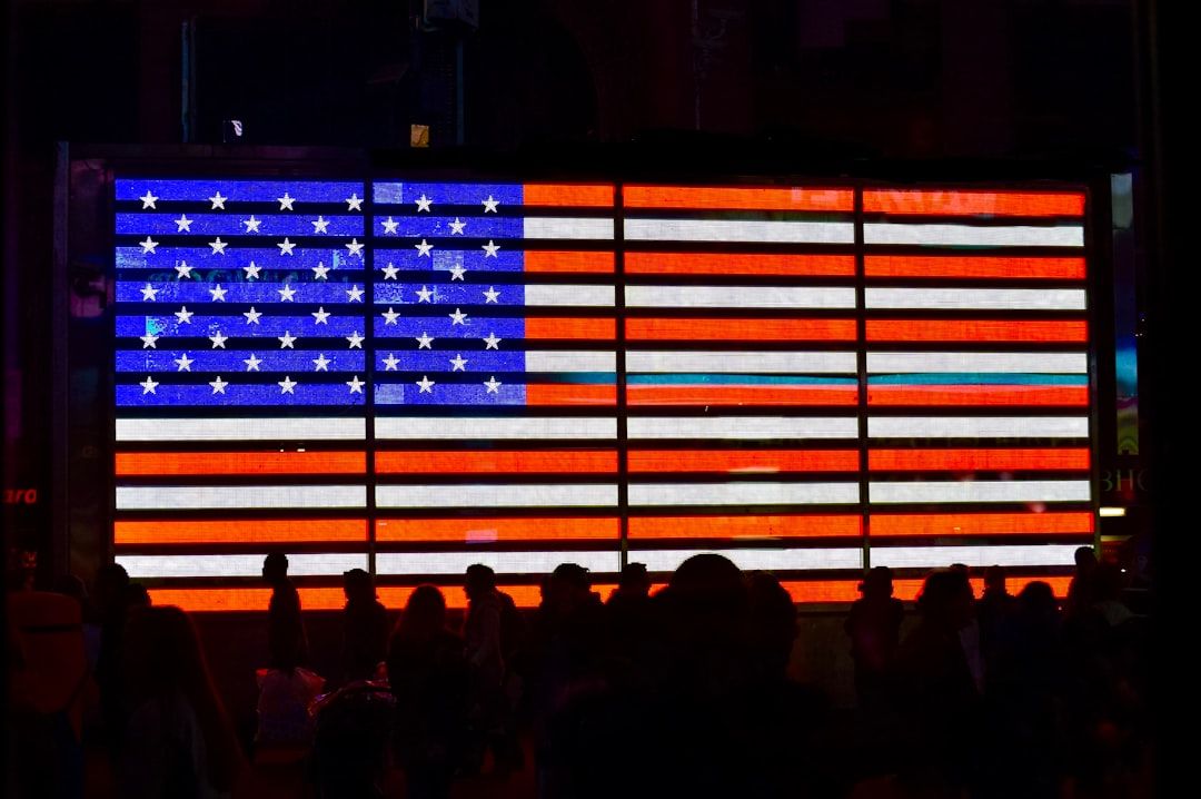 The image depicts a large illuminated display of the American flag in a busy urban setting, with silhouettes of people walking in the foreground. The vibrant colors of the flag contrast against the darkness of the surrounding environment.