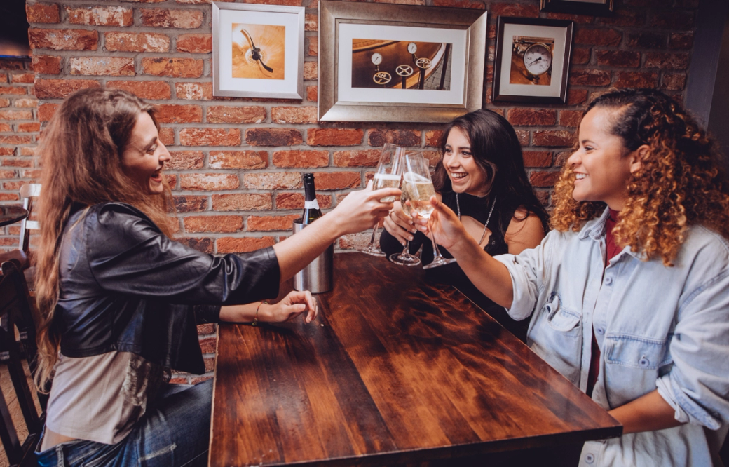 Three women are seated at a wooden table in a cozy brick-walled venue, engaging in a toast with glasses of sparkling wine. The atmosphere appears lively and celebratory, with framed artworks hanging on the walls.