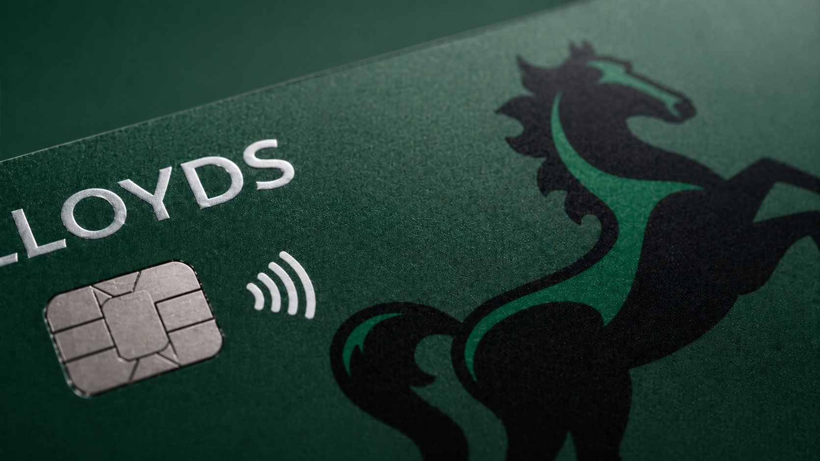 The image features a close-up view of a debit or credit card, highlighting its dark green surface and embossed elements. The card displays the name 'Lloyds' alongside a contactless payment symbol.